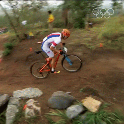 A Man Is Riding A Bike On A Dirt Trail With The Olympic Rings In The Background GIF