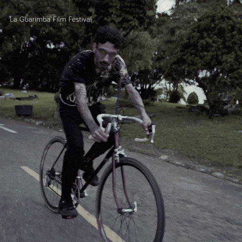 A Man Is Riding A Bike On A Road With La Guarimba Film Festival Written On The Bottom Right GIF