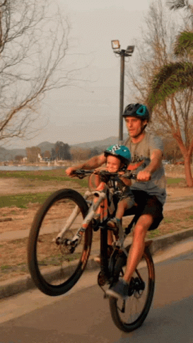 A Man Is Riding A Bike With A Child In A Helmet On The Back GIF