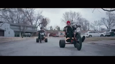 A Man Is Riding A Tricycle Down A Street Next To A Man Riding A Bike . GIF