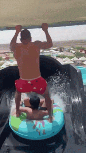 A Man Is Riding A Tube Down A Water Slide While Another Man Stands On Top Of It . GIF