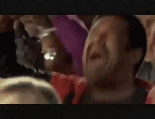 A Man Is Screaming In A Crowd Of People While Holding His Arm In The Air . GIF