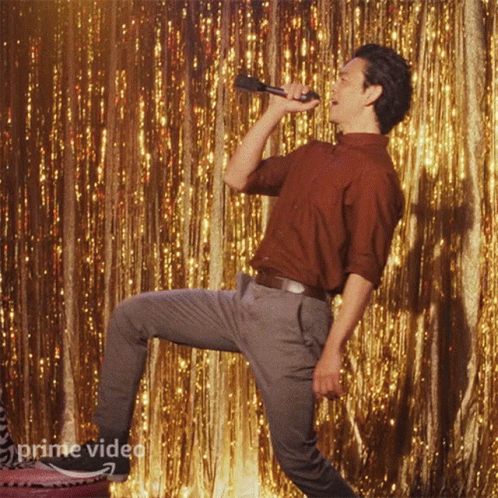 A Man Is Singing Into A Microphone In Front Of A Gold Curtain That Says Prime Video On It GIF