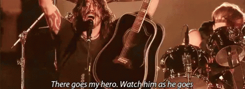 A Man Is Singing Into A Microphone While Holding A Guitar In Front Of A Drum Set . GIF