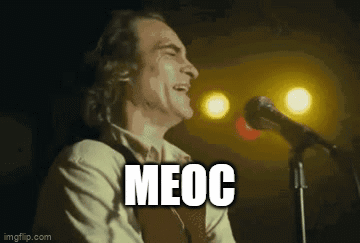 A Man Is Singing Into A Microphone With The Word Meoc Written On The Screen Behind Him . GIF