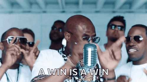 A Man Is Singing Into A Microphone With The Words " A Win Is A Win " Behind Him GIF