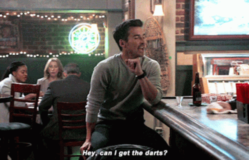 A Man Is Sitting At A Bar Talking To Another Man Who Is Asking If He Can Get The Darts GIF