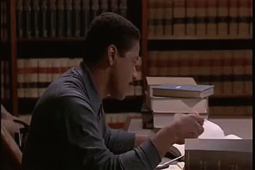 A Man Is Sitting At A Desk Reading A Book And Eating A Sandwich In A Library . GIF