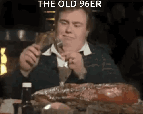 A Man Is Sitting At A Table Eating A Large Piece Of Meat While Holding A Fork . GIF