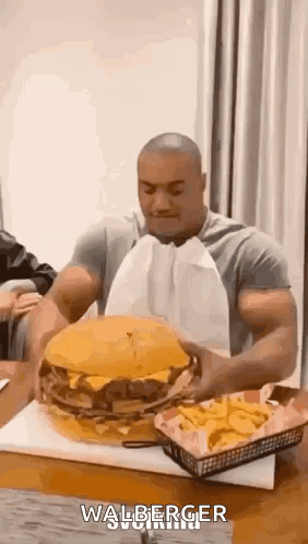A Man Is Sitting At A Table Eating A Very Large Hamburger And French Fries . GIF