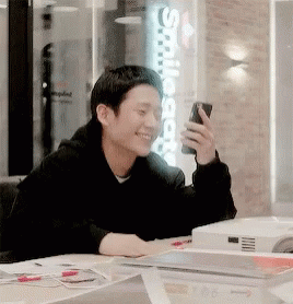 A Man Is Sitting At A Table Holding A Cell Phone And Smiling GIF