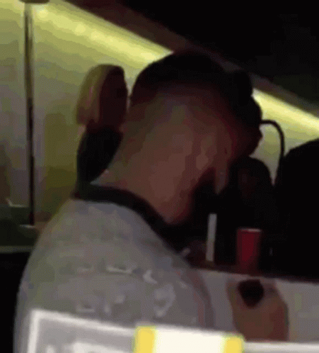 A Man Is Sitting At A Table In A Dark Room With A Red Cup In His Hand . GIF