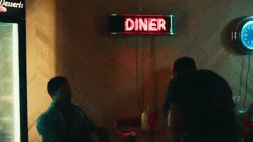 A Man Is Sitting At A Table In A Diner With A Neon Sign Above Him . GIF