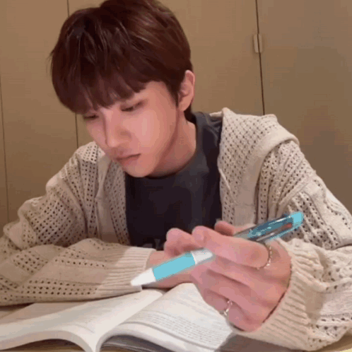 A Man Is Sitting At A Table Reading A Book And Holding A Pen GIF