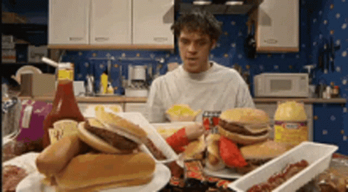 A Man Is Sitting At A Table With A Bunch Of Food Including Hamburgers And Hot Dogs .. GIF