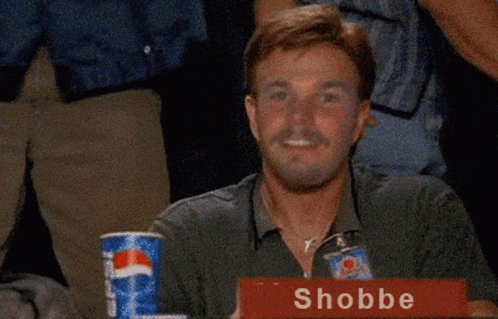 A Man Is Sitting At A Table With A Sign That Says Shobbe On It GIF