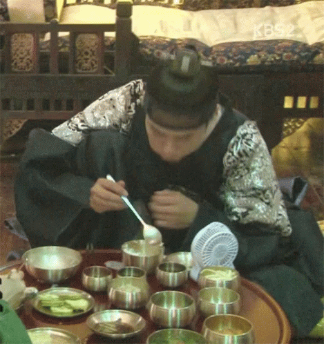 A Man Is Sitting At A Table With Bowls Of Food And A Fan And Eating With A Spoon And Fork GIF