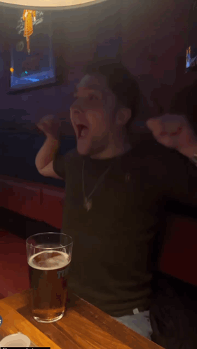 A Man Is Sitting At A Table With His Arms In The Air And A Glass Of Beer In Front Of Him That Says " The " GIF
