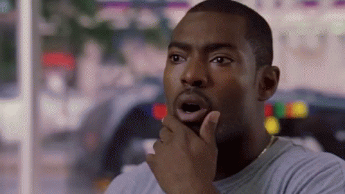A Man Is Sitting At A Table With His Hand On His Chin And A Surprised Look On His Face . GIF