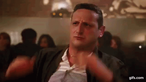 A Man Is Sitting At A Table With His Hands On His Chest In A Crowded Room . GIF