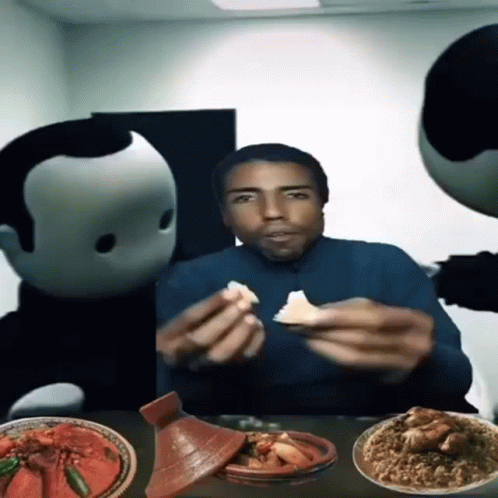A Man Is Sitting At A Table With Plates Of Food And Two Mascots Behind Him GIF