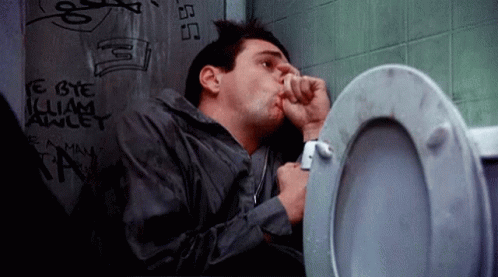 A Man Is Sitting In A Bathroom Next To A Toilet With Graffiti On The Wall Behind Him . GIF