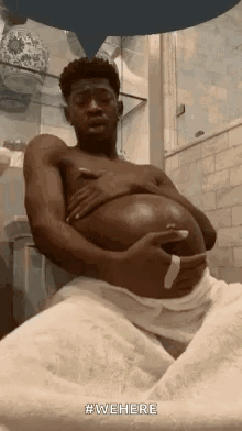 A Man Is Sitting In A Bathroom With A Towel Around His Waist And Holding His Belly . GIF