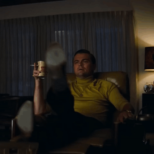 A Man Is Sitting In A Chair Holding A Can Of Potato Chips And Smoking A Cigarette GIF