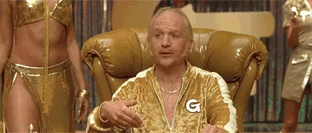 A Man Is Sitting In A Chair Wearing A Gold Jacket With A G On It . GIF