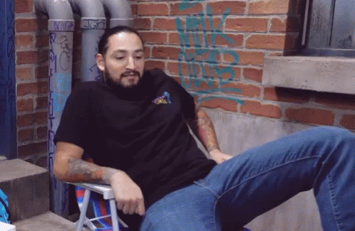 A Man Is Sitting In A Chair With A Brick Wall Behind Him With Graffiti On It That Says ' Rick ' GIF