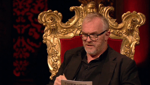 A Man Is Sitting In A Chair With A Piece Of Paper That Says Taskmaster On It GIF