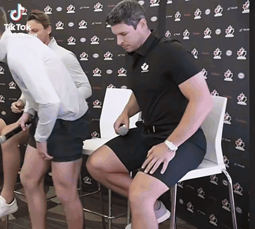 A Man Is Sitting In A Chair With His Legs Crossed While Another Man Stands Behind Him Holding A Microphone .. GIF