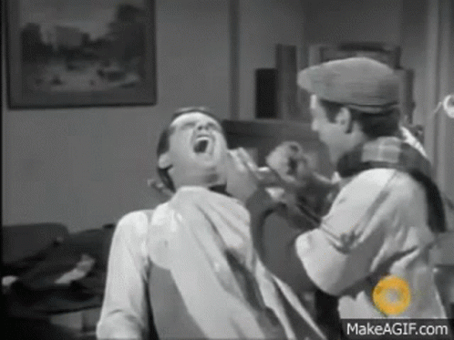 A Man Is Sitting In A Chair With His Mouth Open While Another Man Shaves His Beard . GIF