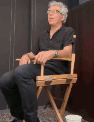 A Man Is Sitting In A Director 'S Chair With His Legs Crossed GIF