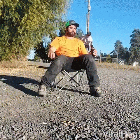 A Man Is Sitting In A Folding Chair On The Side Of A Road Holding A Bottle Of Beer . GIF