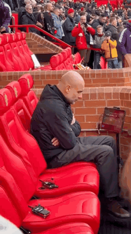 A Man Is Sitting In A Row Of Red Seats In A Stadium With His Arms Crossed . GIF