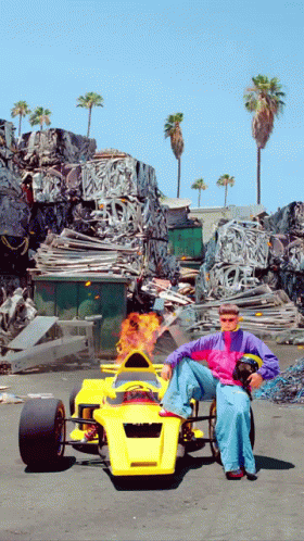 A Man Is Sitting In A Yellow Race Car With Flames Coming Out Of The Steering Wheel GIF