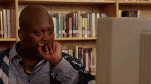 A Man Is Sitting In Front Of A Computer In A Library With His Hand On His Chin . GIF