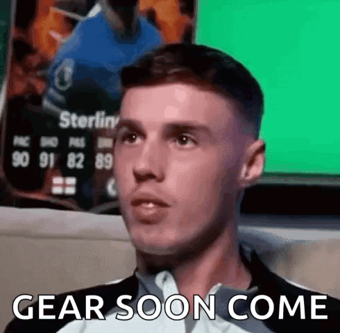 A Man Is Sitting In Front Of A Green Screen With The Words `` Gear Soon Come '' Written On It . GIF