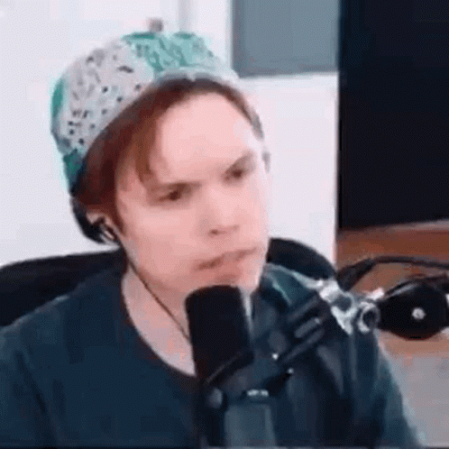 A Man Is Sitting In Front Of A Microphone Wearing A Hat And Headphones . GIF