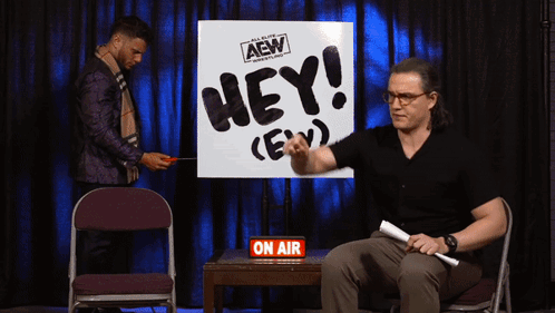 A Man Is Sitting In Front Of A Sign That Says Hey GIF
