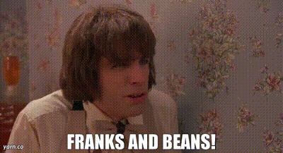 A Man Is Sitting In Front Of A Wall With Flowers On It And Says `` Franks And Beans '' . GIF