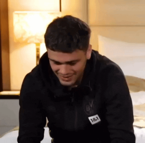 A Man Is Sitting On A Bed Wearing A Black Jacket With The Number 18 On It . GIF