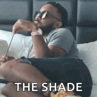A Man Is Sitting On A Bed Wearing Sunglasses And Holding A Pair Of Scissors . GIF