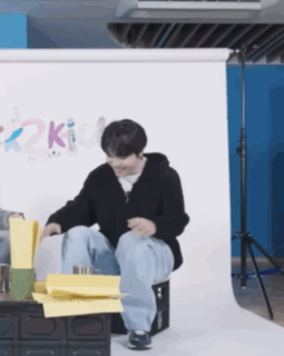 A Man Is Sitting On A Box With His Legs Crossed In Front Of A Wall With The Word Kit On It GIF