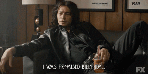 A Man Is Sitting On A Couch And Says " I Was Promised Billy Idol " While Holding A Cup GIF