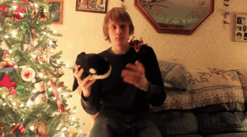 A Man Is Sitting On A Couch Holding A Stuffed Animal In Front Of A Christmas Tree GIF