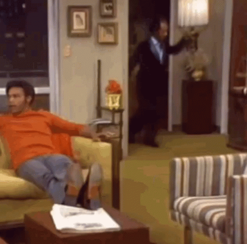 A Man Is Sitting On A Couch In A Living Room While A Man In A Suit Is Standing In The Doorway . GIF