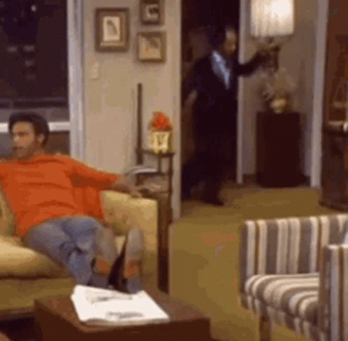 A Man Is Sitting On A Couch In A Living Room While A Man In A Suit Stands In The Doorway . GIF