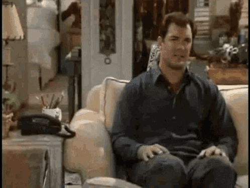 A Man Is Sitting On A Couch In A Living Room With A Telephone On The Table . GIF
